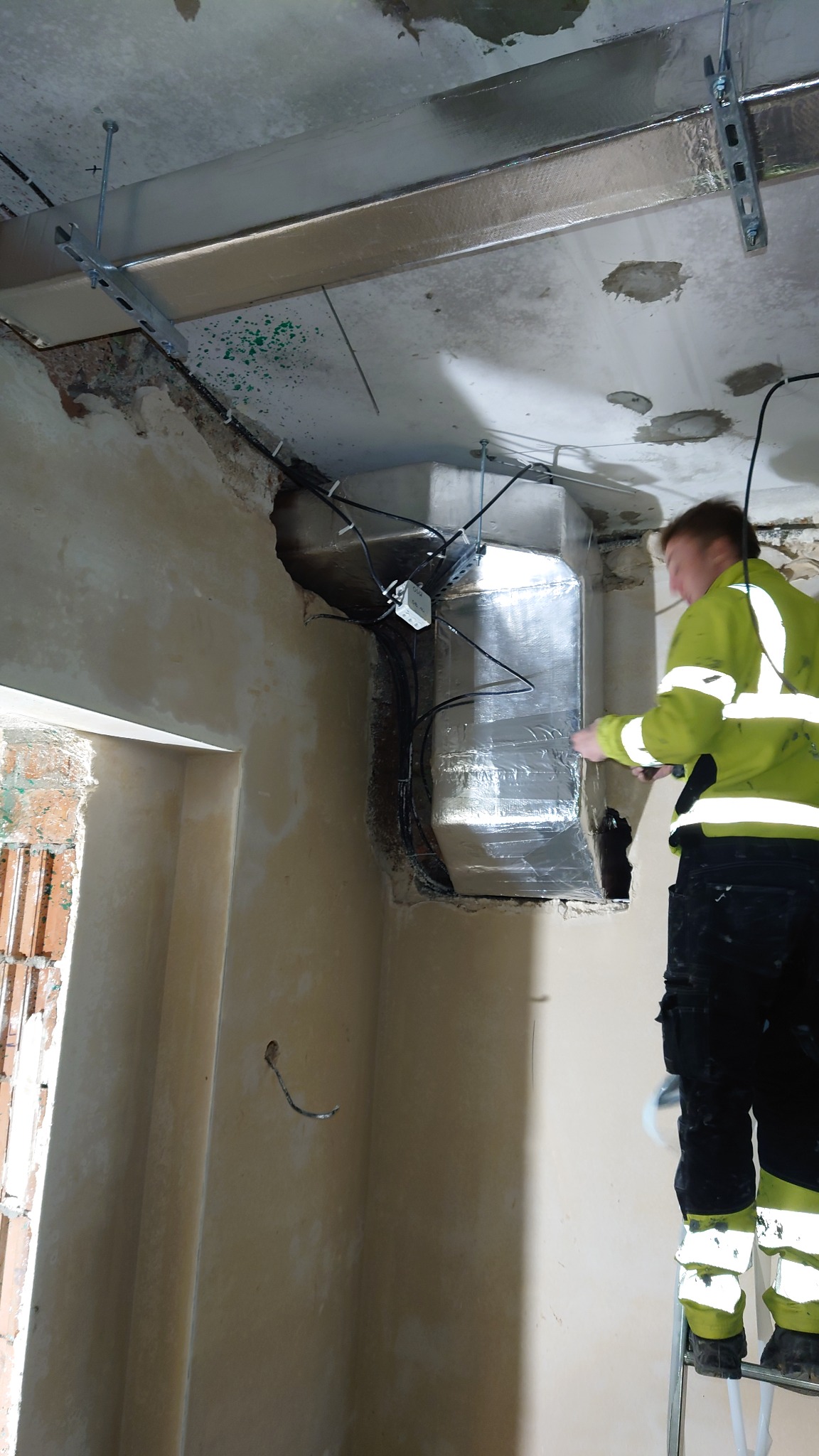 Pracownik w odblaskowej kurtce na drabinie, instaluje prostokątny kanał wentylacyjny w surowym wnętrzu, widoczne przewody i elementy mocujące do sufitu.