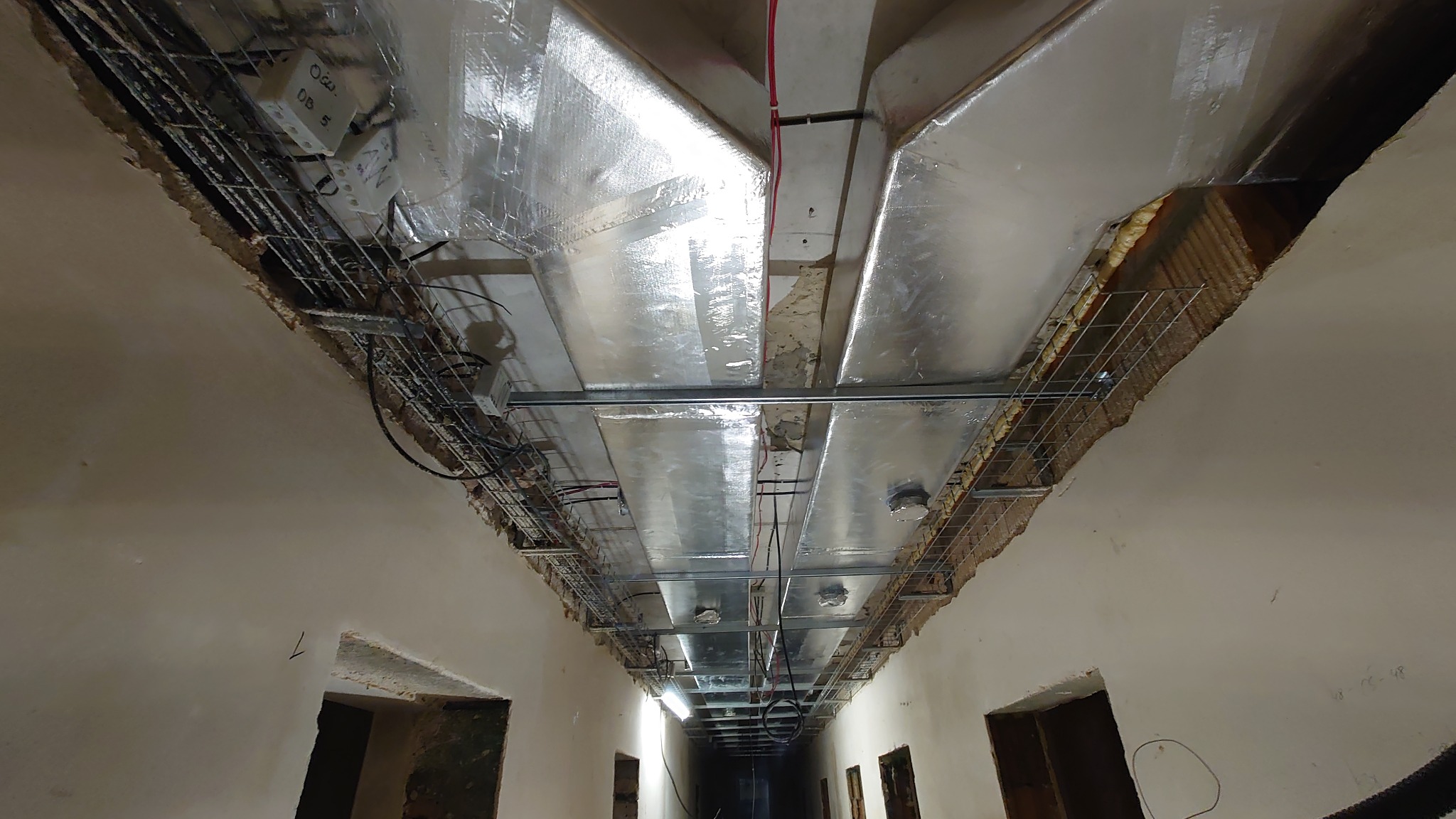 Widok z dołu na system kanałów wentylacyjnych, z widoczną izolacją termiczną, metalowymi korytami kablowymi i przewodami, w korytarzu budynku w trakcie remontu.