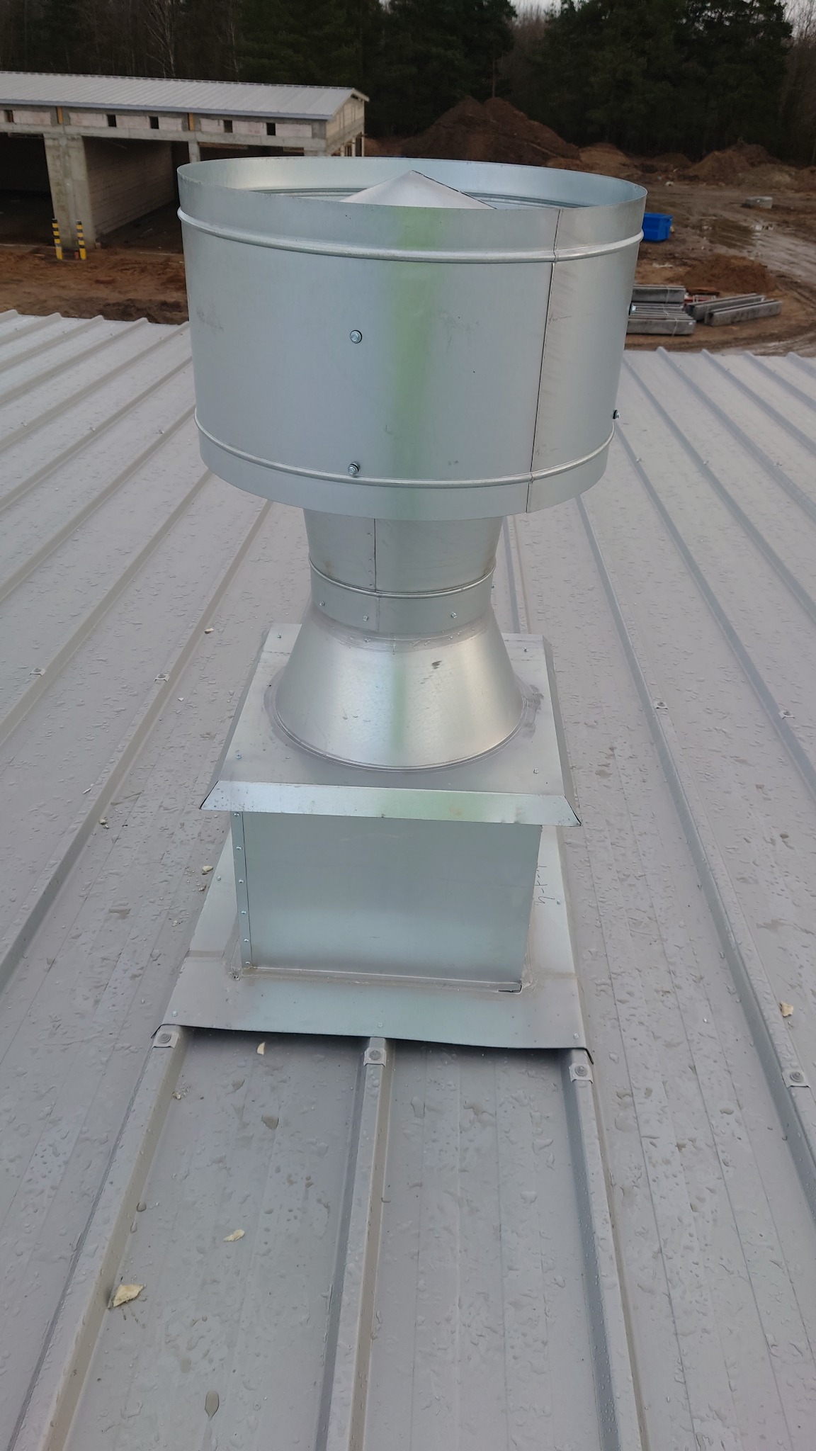 Metalowy wywiewnik na dachu z blachy trapezowej, widoczne ślady po deszczu, w tle budynek w stanie surowym otwartym i hałda ziemi.