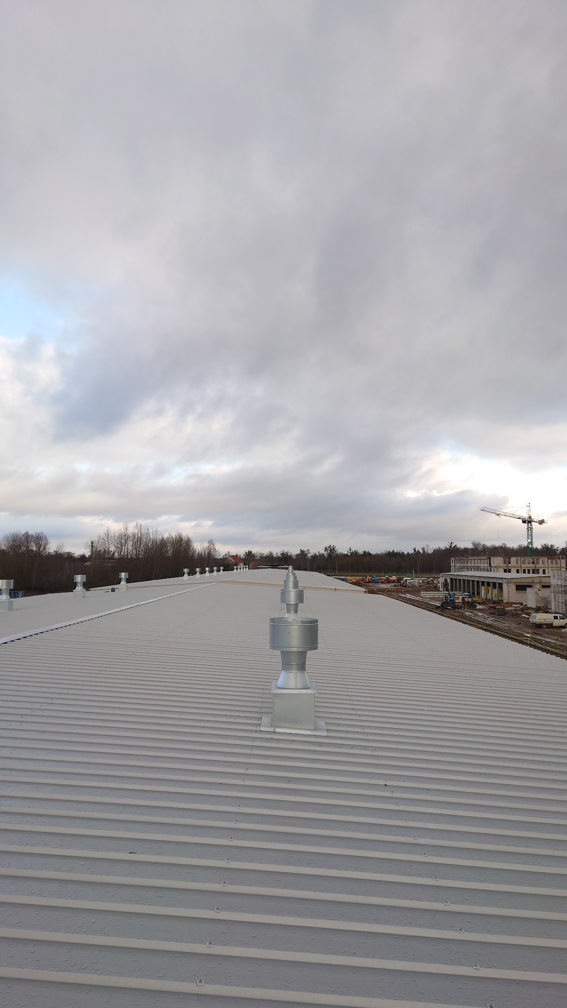 Szary dach budynku komercyjnego z zamontowanymi srebrnymi kominami wentylacyjnymi i elementami rekuperacji, w tle budowa i pochmurne niebo.