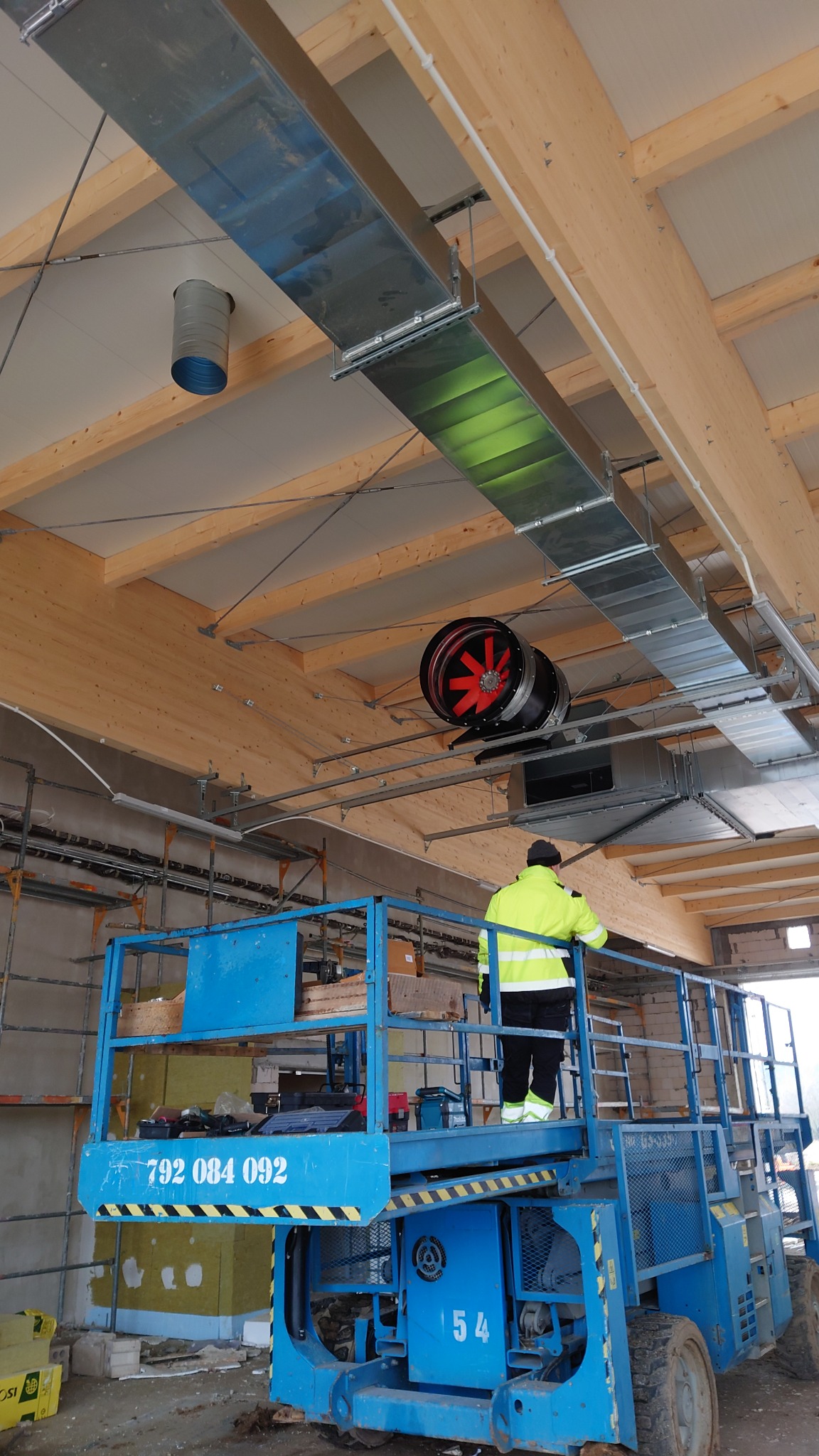 Pracownik w żółtej kurtce na podnośniku hydraulicznym montuje system kanałów wentylacyjnych z czarnym wentylatorem z czerwonymi łopatkami w jasnym pomieszczeniu z drewnianą konstrukcją dachu.