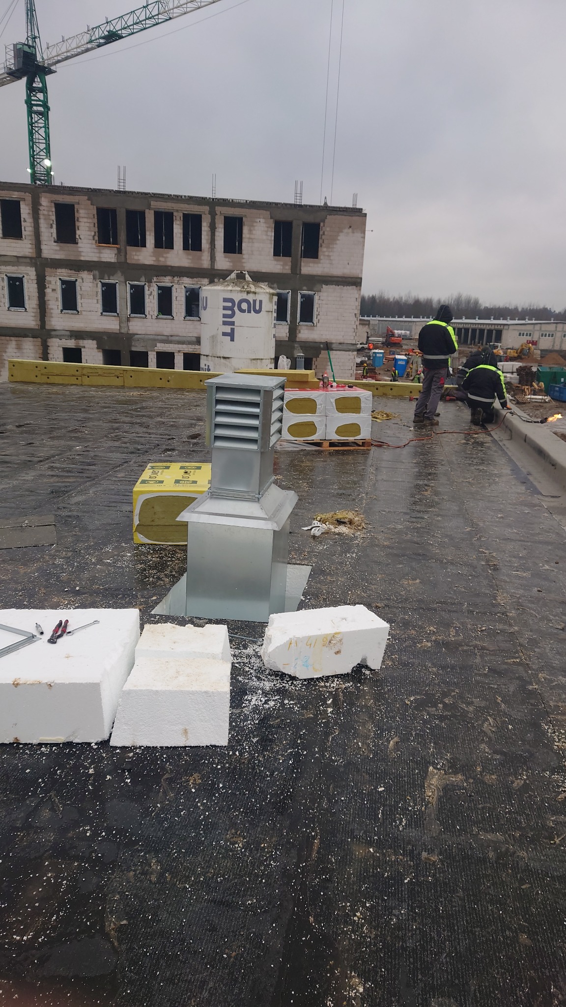 Metalowa czerpnia powietrza na dachu budynku w trakcie instalacji, widoczny izolacja, styropian i pracownicy przy pracy.