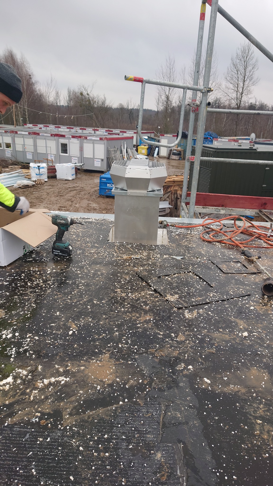 Montaż metalowego komina wentylacyjnego na dachu pokrytym czarną papą z białymi plamkami, w tle widoczne baraki budowlane i rusztowanie, w kadrze wiertarka akumulatorowa i fragment osoby...