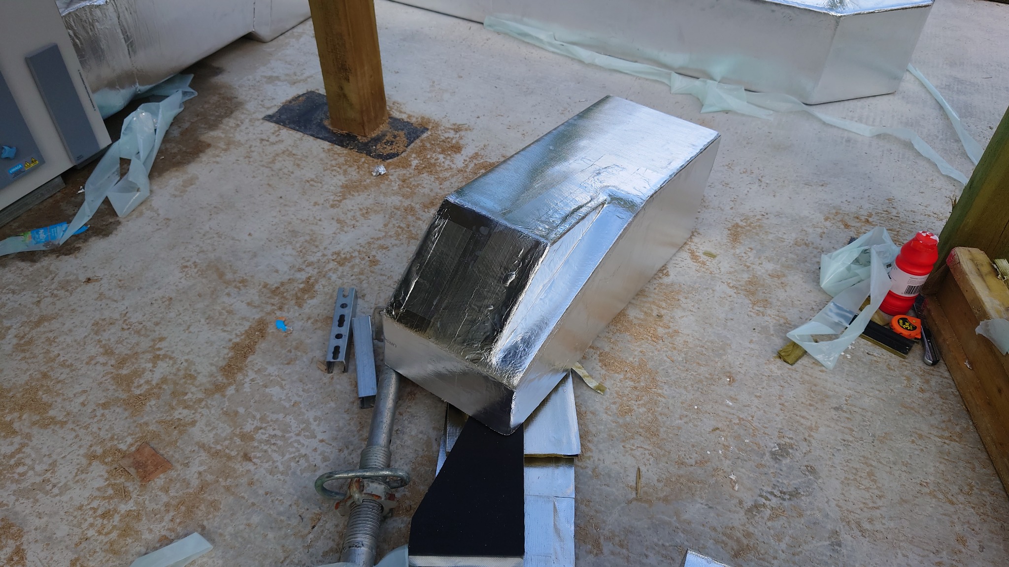 Fragment izolowanego, srebrną folią kanału wentylacyjnego, leżącego na podłodze w trakcie montażu, widoczne narzędzia i elementy montażowe.
