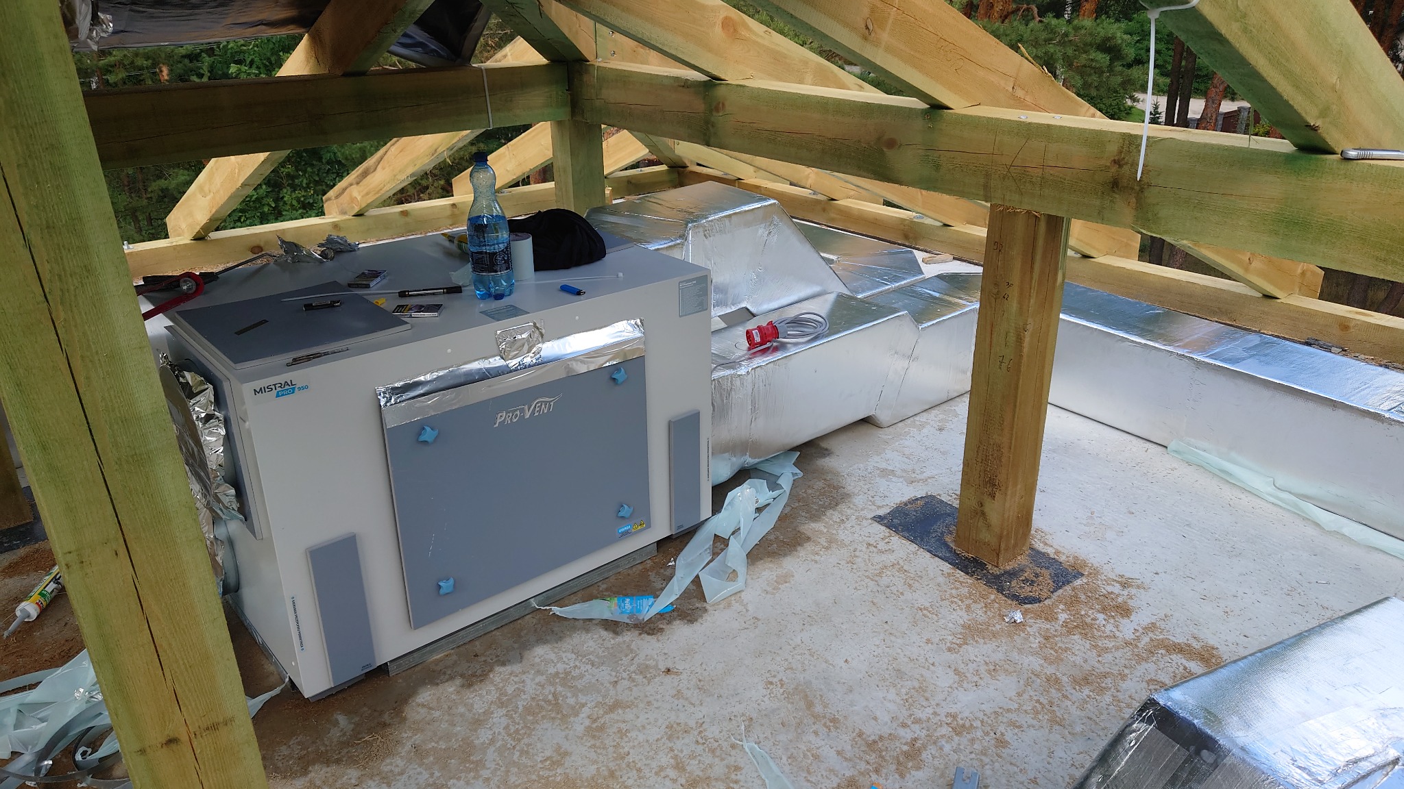 Centrala wentylacyjna Mistral Pro 950 z kanałami wentylacyjnymi w srebrnej izolacji, zamontowana w surowym stanie poddasza z widoczną drewnianą konstrukcją dachu.