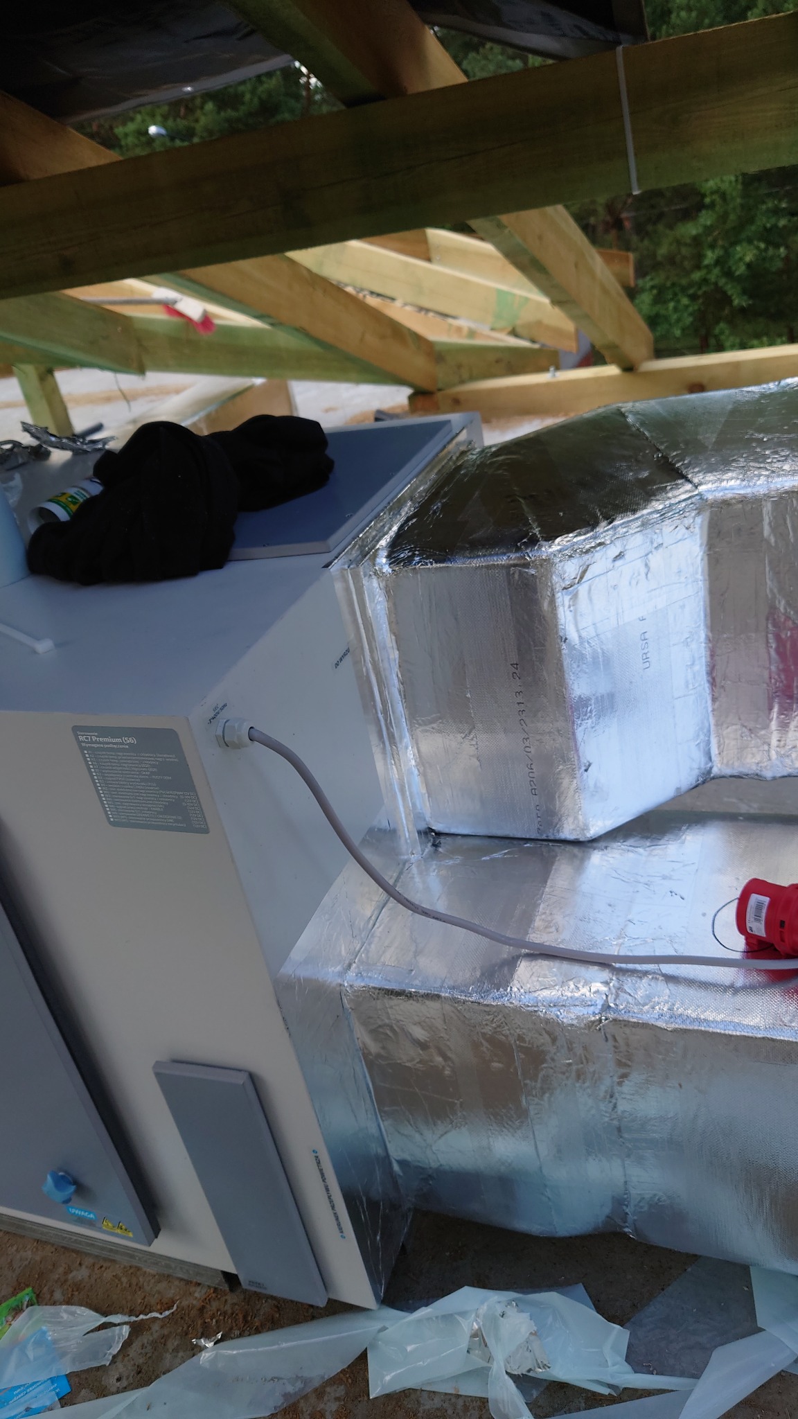 Elementy systemu wentylacyjnego z izolacją termiczną z folii aluminiowej, widoczne poddasze z drewnianą konstrukcją dachu i czarna bluza na urządzeniu.