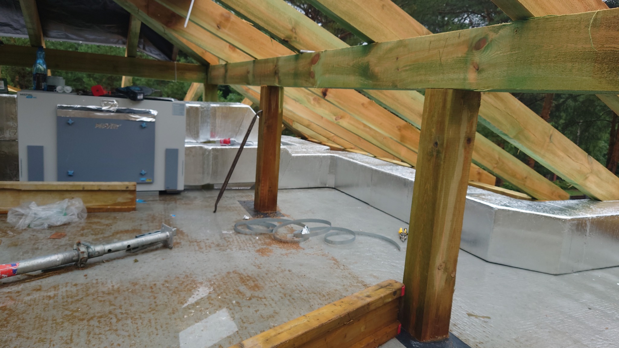 Wnętrze poddasza w trakcie prac: drewniana konstrukcja dachu, izolacja ścian folią aluminiową, widoczny centralny element instalacji wentylacyjnej z nazwą producenta, narzędzia i elementy montażowe...