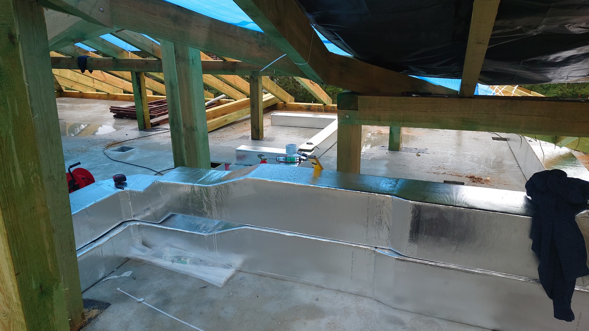 Wnętrze budynku w trakcie budowy z widocznymi kanałami wentylacyjnymi z izolacją aluminiową, narzędziami i elementami konstrukcyjnymi dachu.