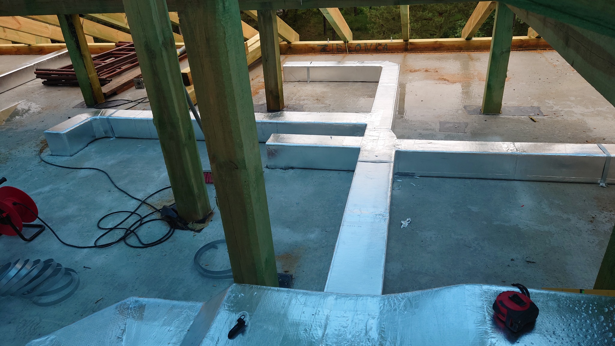 Montaż izolowanych kanałów wentylacyjnych w surowym stanie budynku w Mińsku Mazowieckim, widoczne drewniane elementy konstrukcyjne dachu i narzędzia pracy.