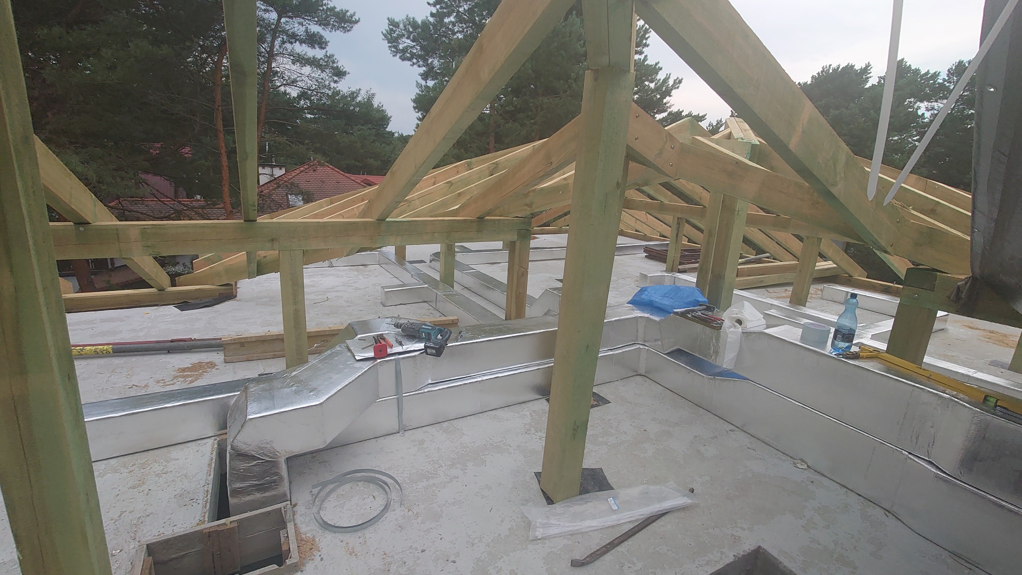 Szare, metalowe kanały wentylacyjne ułożone na betonowej podłodze pod drewnianą konstrukcją dachu, widoczne narzędzia i materiały budowlane.