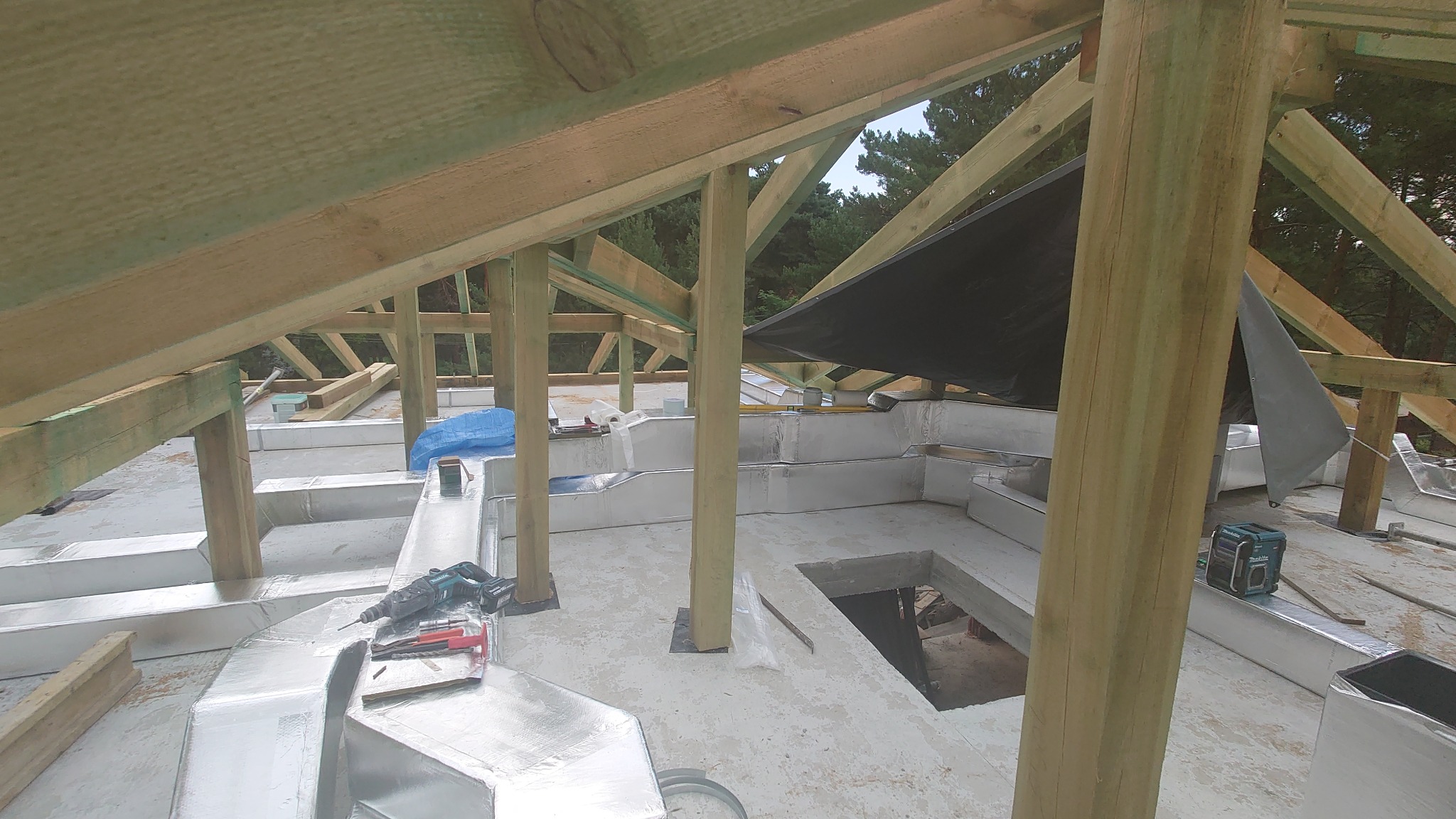 Widok z góry na montaż systemu kanałów wentylacyjnych w przestrzeni poddasza, z widoczną konstrukcją drewnianą dachu i narzędziami pracy.