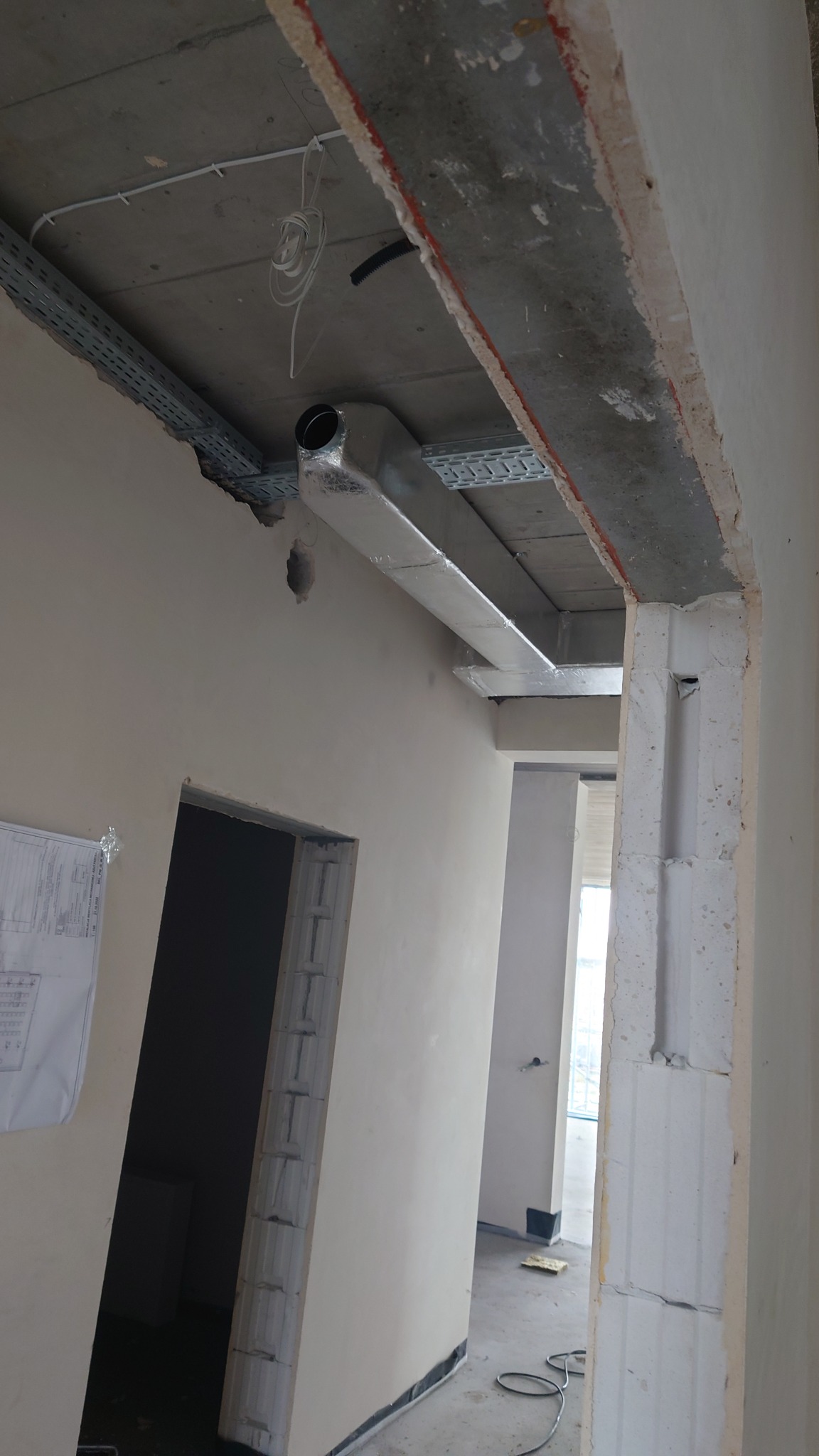 Instalacja kanału wentylacyjnego z izolacją termiczną pod sufitem w surowym wnętrzu budynku. Widoczne kable, profile montażowe i otwór w ścianie. W tle widoczny niezakończony stan surowy.