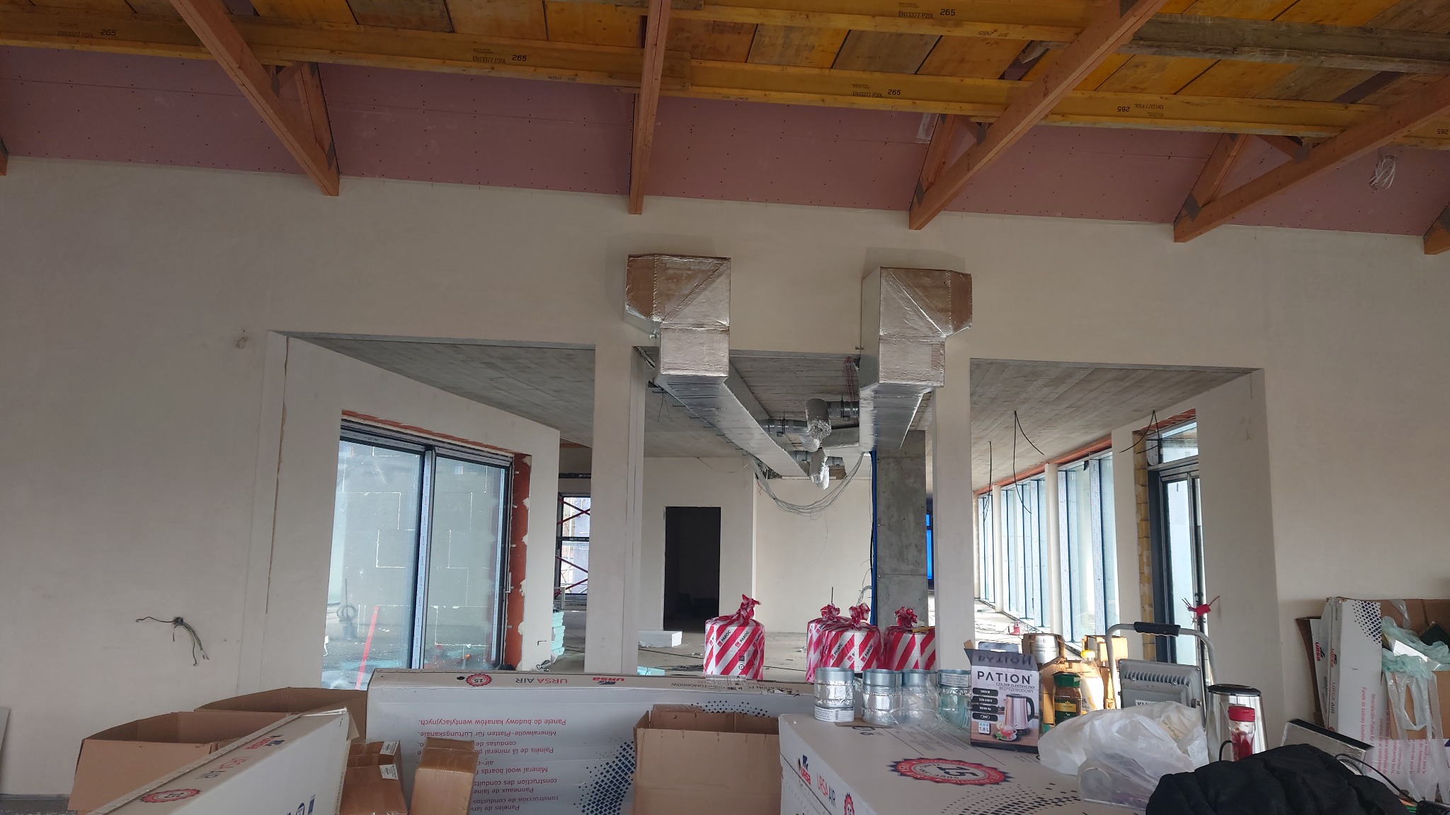 Montaż instalacji wentylacyjnej w surowym budynku komercyjnym, widoczne kanały wentylacyjne pod sufitem, materiały budowlane i okna w tle.