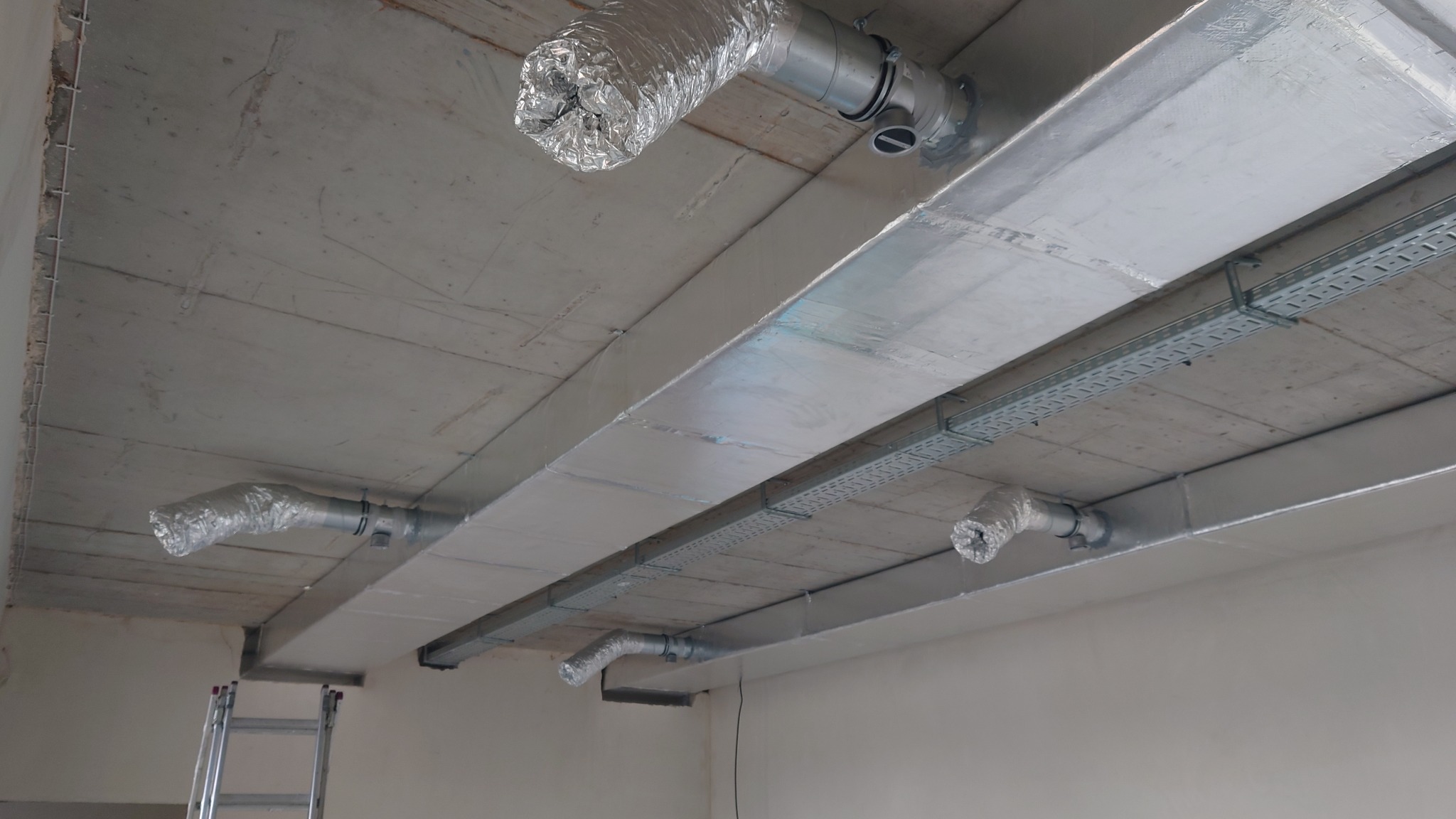 Widok z dołu na sufit z zamontowanymi kanałami wentylacyjnymi, rurami w izolacji aluminiowej i korytkami kablowymi, drabina oparta o ścianę w rogu pomieszczenia.