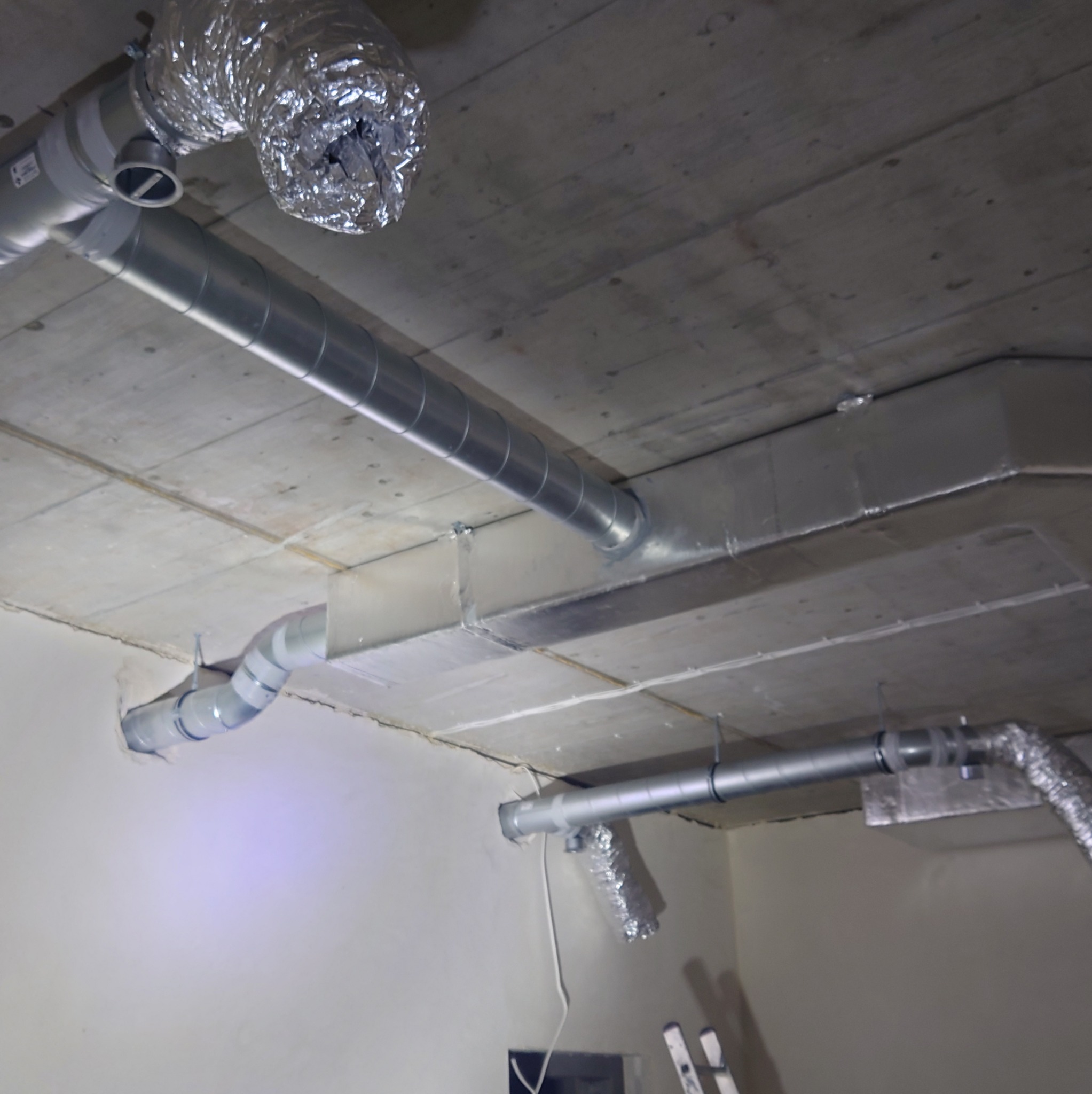 System kanałów wentylacyjnych z rur spiro i elastycznych przewodów aluminiowych zamontowany pod betonowym stropem w surowym wnętrzu, widoczne mocowania i fragment drabiny.