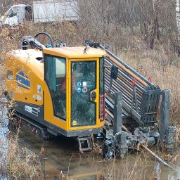 Arek od Koparek Arkadiusz Przewodek - Żółta maszyna do przewiertów sterowanych zanurzona w wodzie na zewnątrz, z rurami w uchwycie. W tle widoczny biały samochód dostawczy i drzewa.