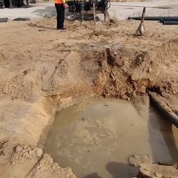 Arek od Koparek Arkadiusz Przewodek - Wykopany dół wypełniony wodą, obok rury i maszyna wiertnicza z pracownikami w odblaskowych kamizelkach. W tle hałda ziemi i las.