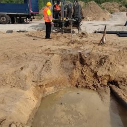 Arek od Koparek Arkadiusz Przewodek - Prace ziemne: operator w żółtej kabinie wiertnicy, obok pracownik w kamizelce. Wykop z wodą, rury i ciężarówka w tle. Dzień, teren budowy.