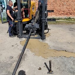 Arek od Koparek Arkadiusz Przewodek - Żółta maszyna do przewiertów sterowanych w trakcie pracy. Widoczni dwaj operatorzy, błoto wokół wiertła i cień osoby robiącej zdjęcie. Ceglany budynek w tle.