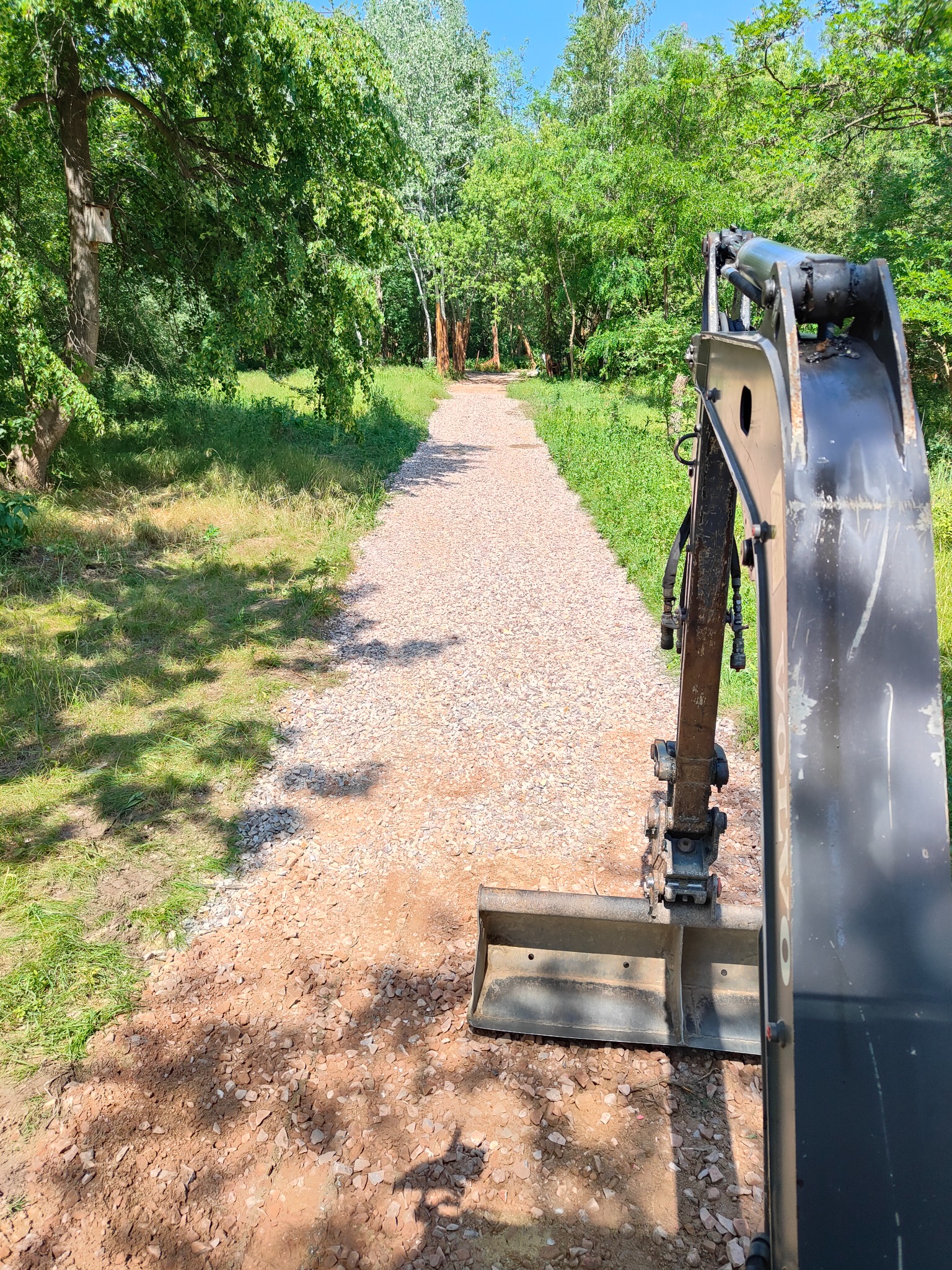 Koparka z łyżką widoczna z bliska na tle nowo utwardzonej, żwirowej ścieżki w parku, otoczonej zielenią.
