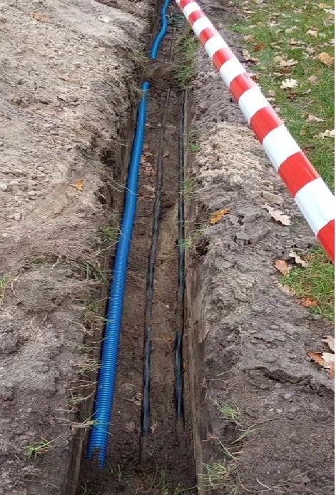 Wykop z ułożonym niebieskim karbowanym przewodem instalacyjnym i czarnymi rurami, zabezpieczony biało-czerwoną taśmą ostrzegawczą.