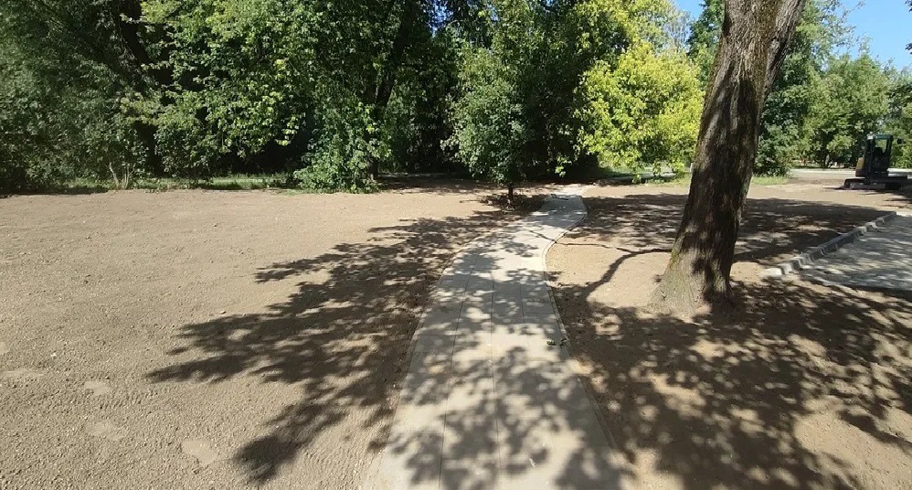 Przygotowany teren pod budowę z ułożonym chodnikiem z kostki betonowej, widoczny cień drzew i maszyna budowlana w tle.