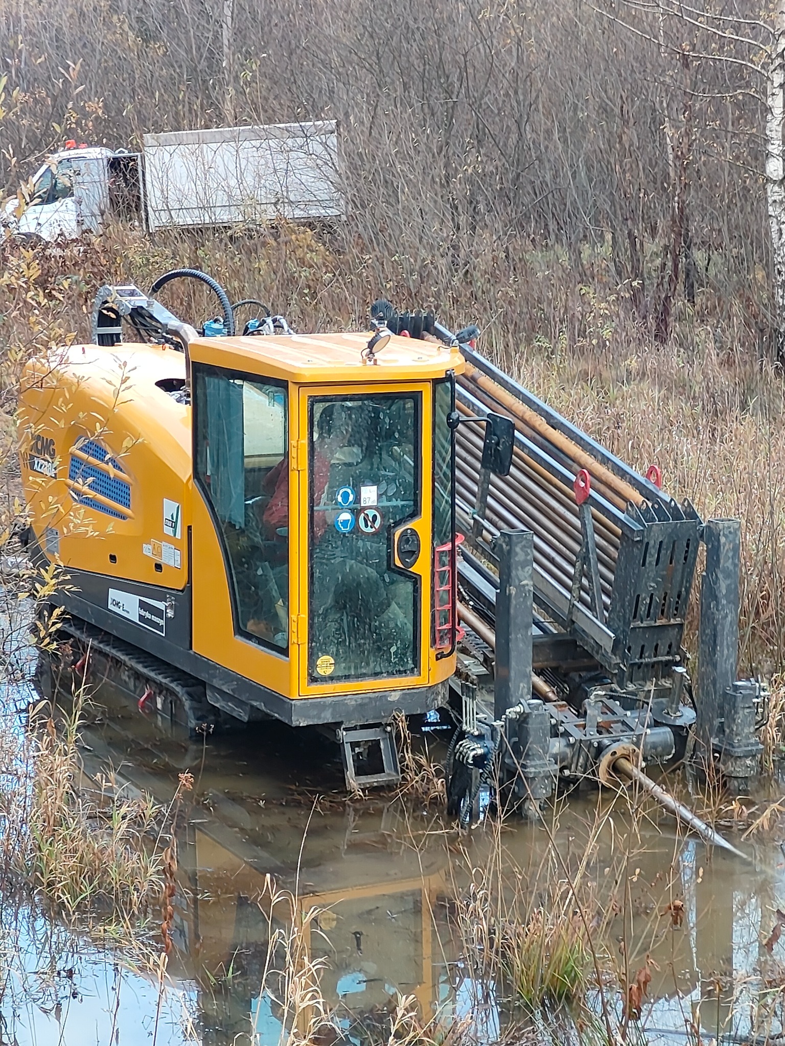 Żółta maszyna do przewiertów sterowanych zanurzona w wodzie na zewnątrz, z rurami w uchwycie. W tle widoczny biały samochód dostawczy i drzewa.