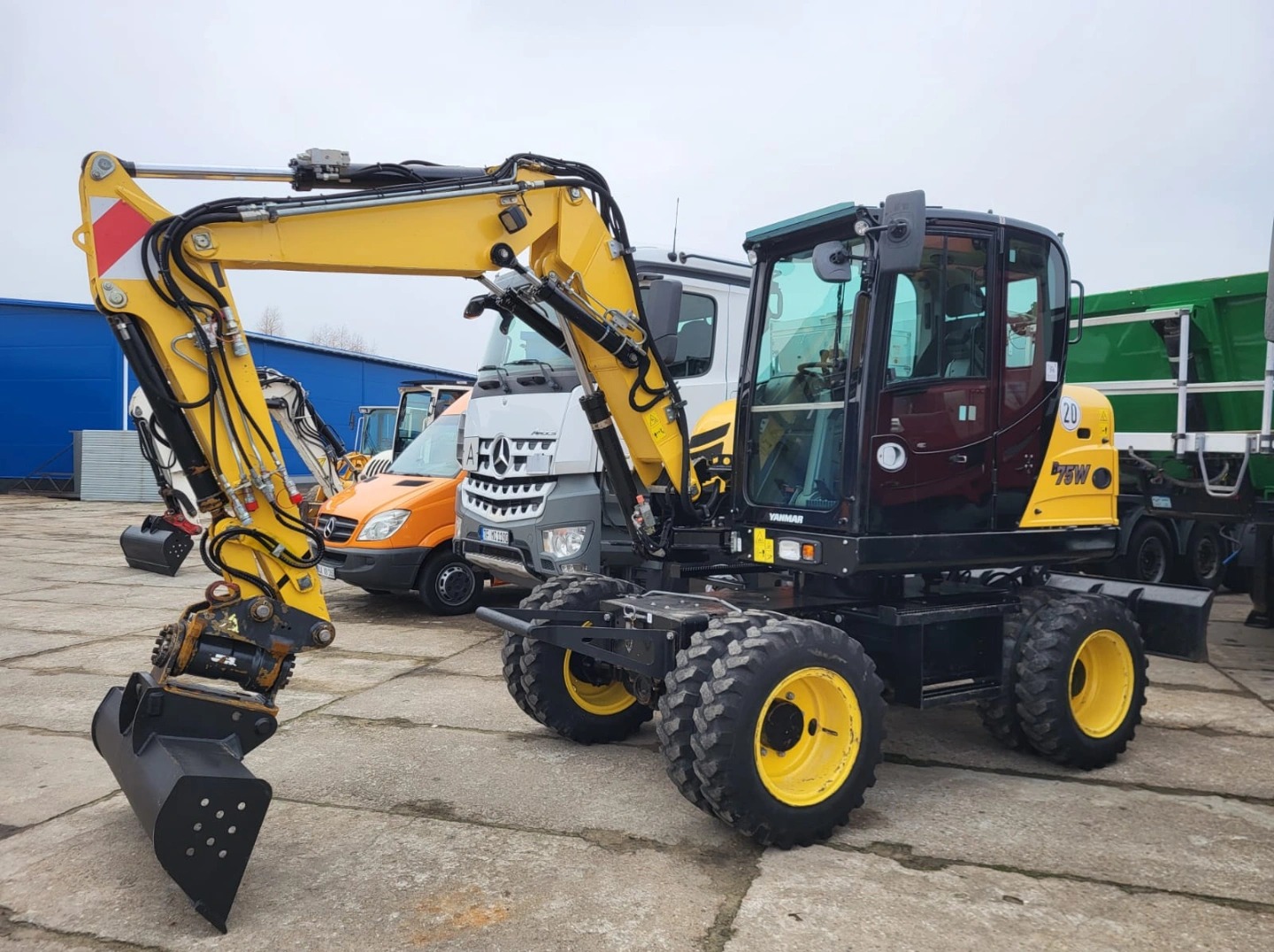 Żółto-czarna koparka kołowa Yanmar 75W z łyżką na betonowym placu, w tle inne pojazdy budowlane i niebieski budynek. Sprzęt gotowy do pracy.