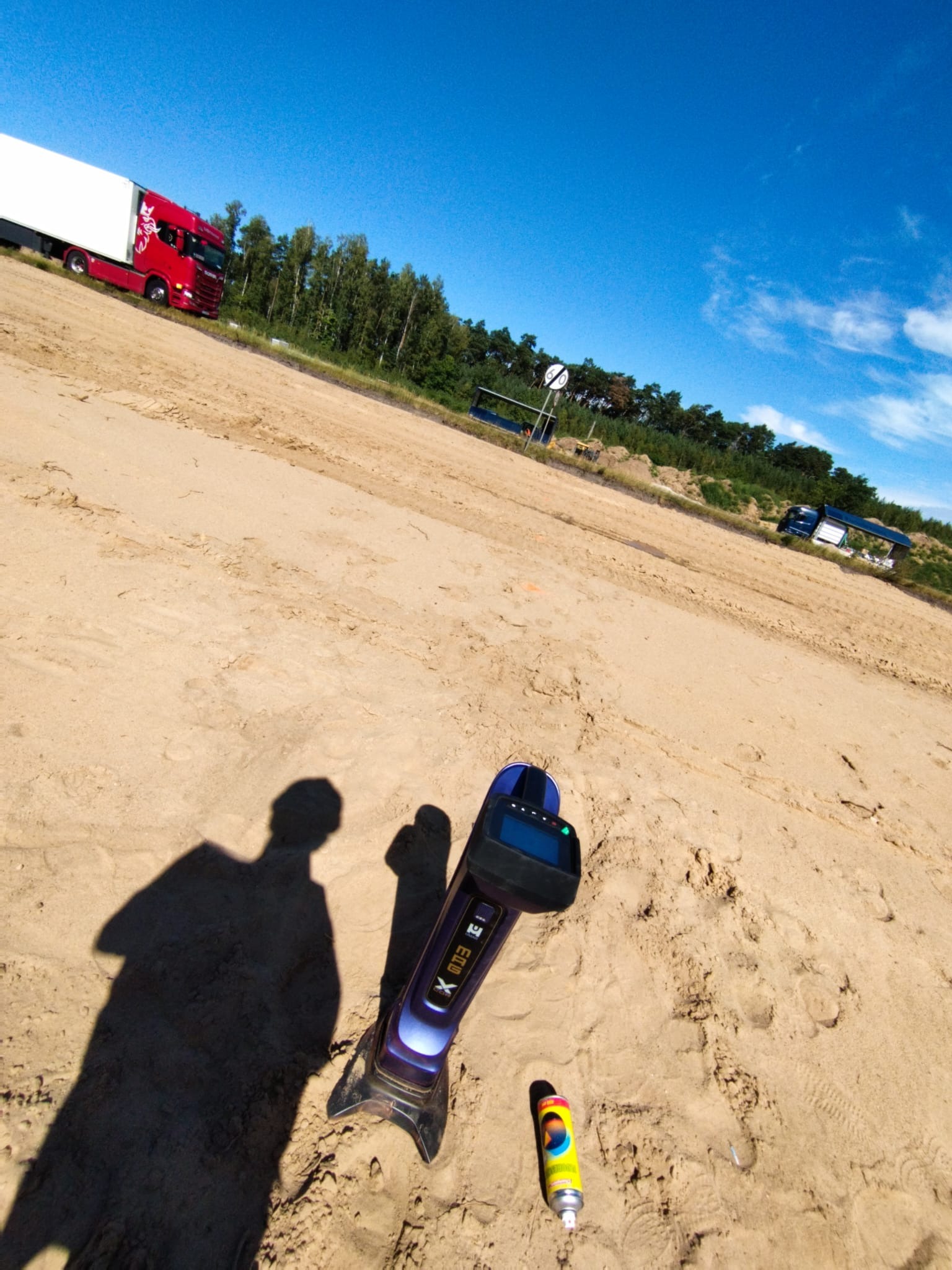 Niwelator gruntu na piaszczystym terenie budowy, cień osoby, ciężarówka w tle, błękitne niebo. Sprzęt pomiarowy w trakcie pracy.