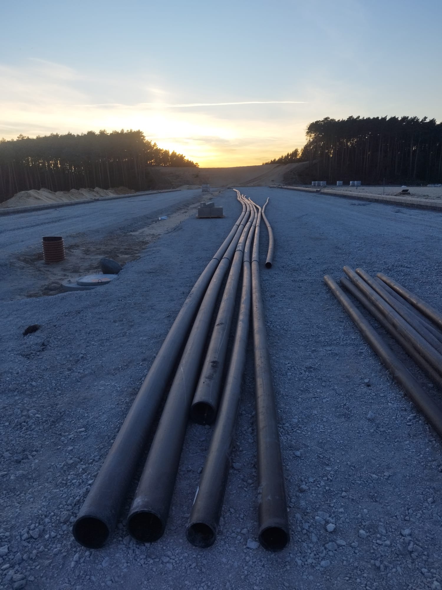 Rury ułożone na żwirowym podłożu wzdłuż drogi, w tle zachodzące słońce i las. Widok na przygotowanie do instalacji podziemnej infrastruktury.