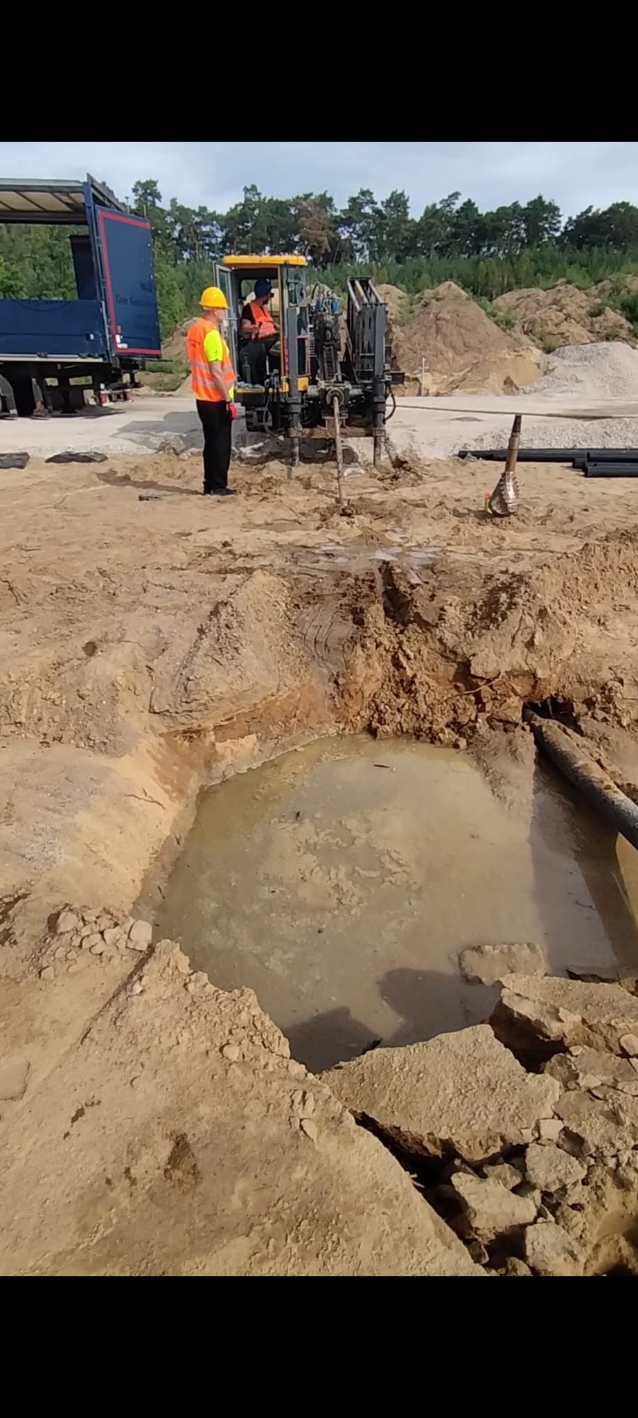 Wykopany dół wypełniony wodą, obok rury i maszyna wiertnicza z pracownikami w odblaskowych kamizelkach. W tle hałda ziemi i las.