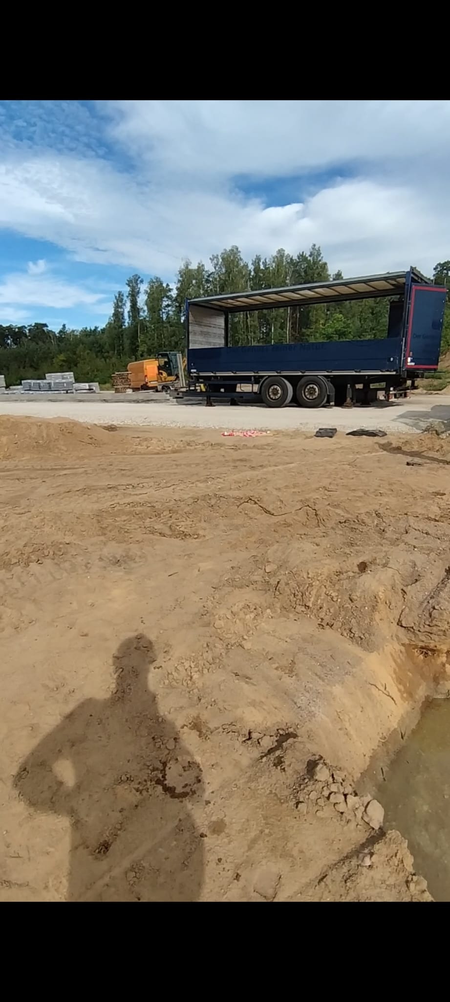 Przygotowany teren pod budowę z widocznym transportem materiałów i koparką w tle, cień osoby w lewym dolnym rogu. Słoneczny dzień, błękitne niebo.