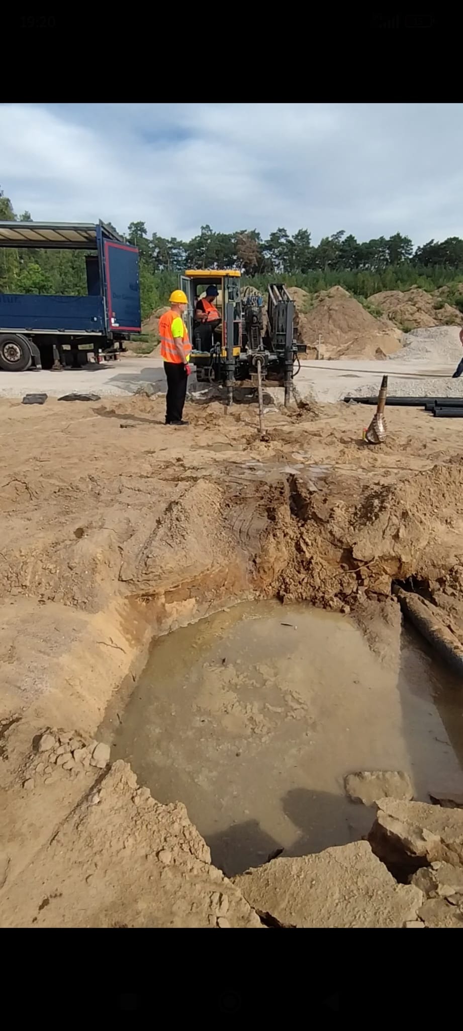 Prace ziemne: operator w żółtej kabinie wiertnicy, obok pracownik w kamizelce. Wykop z wodą, rury i ciężarówka w tle. Dzień, teren budowy.