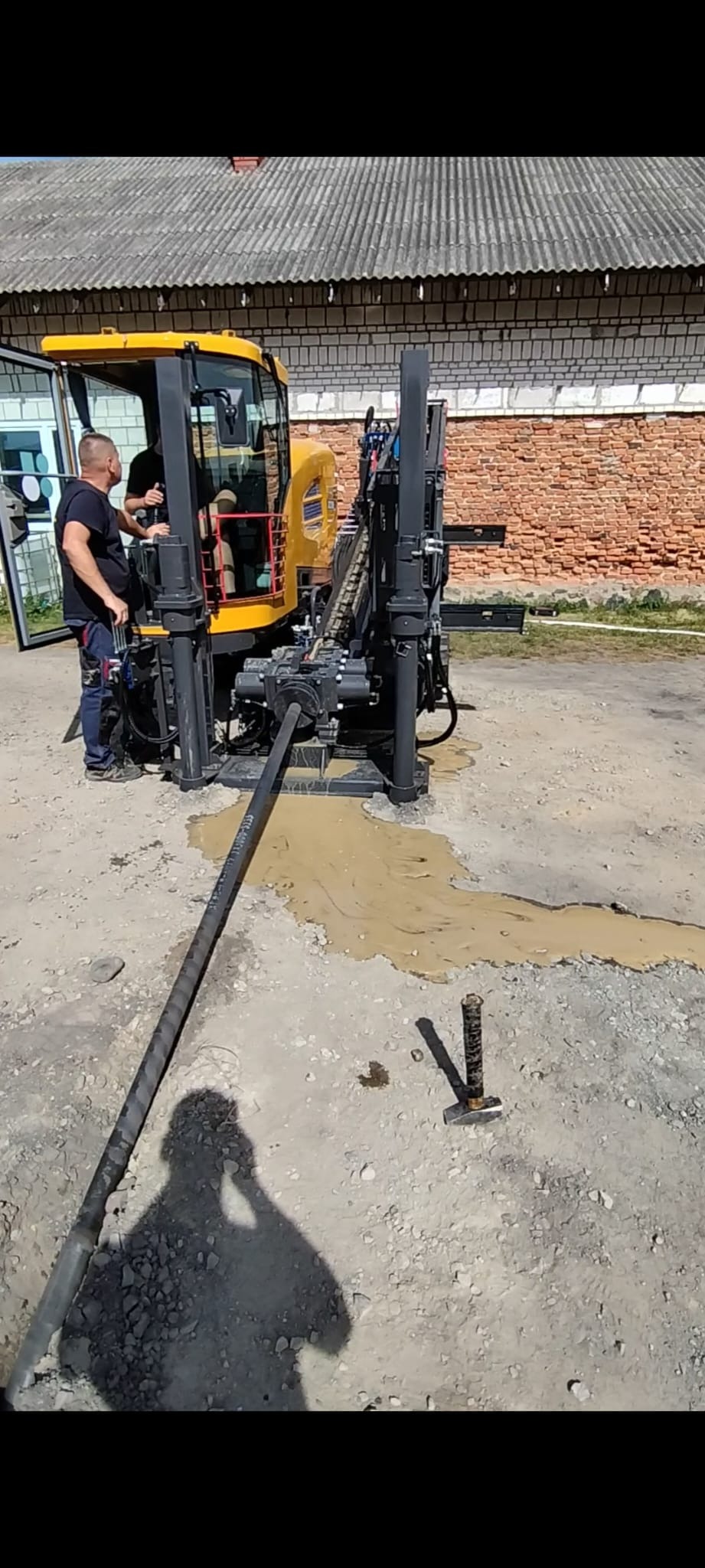 Żółta maszyna do przewiertów sterowanych w trakcie pracy. Widoczni dwaj operatorzy, błoto wokół wiertła i cień osoby robiącej zdjęcie. Ceglany budynek w tle.