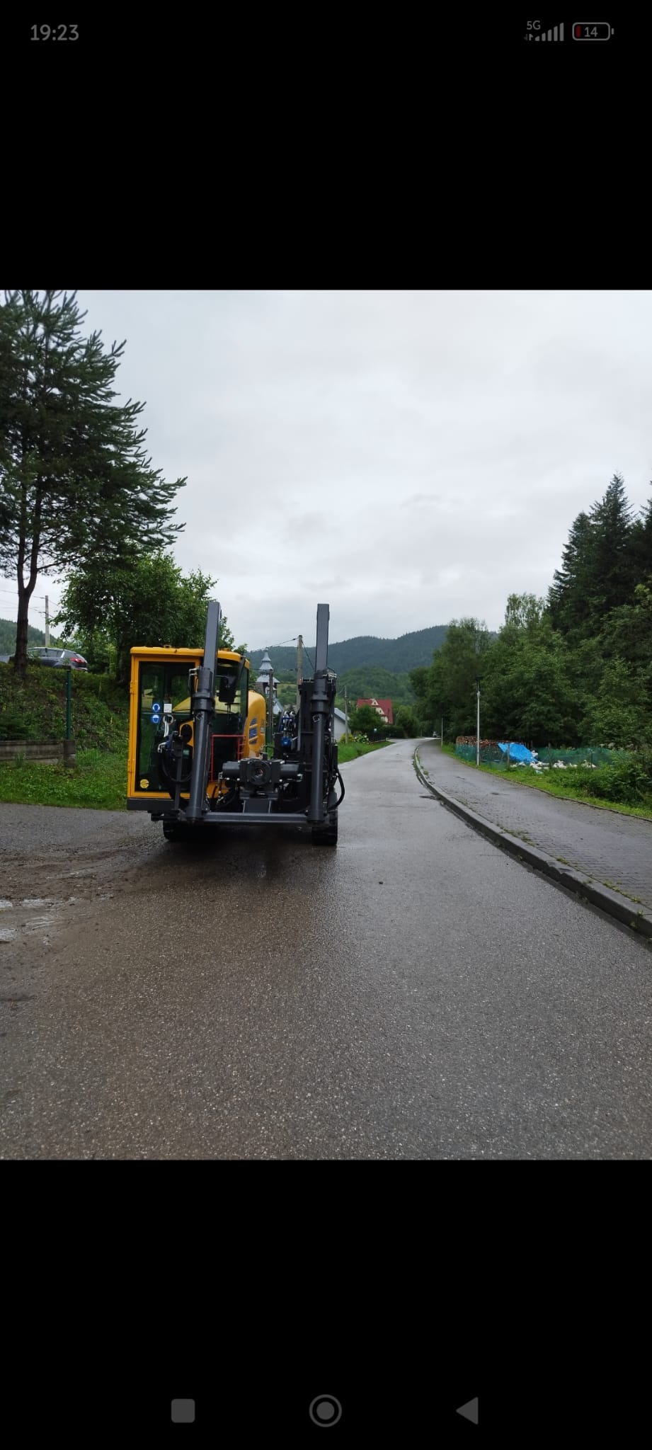 Żółta maszyna do przewiertów sterowanych na mokrej, asfaltowej drodze w otoczeniu zieleni i gór. Widok z tyłu, pochmurne niebo.