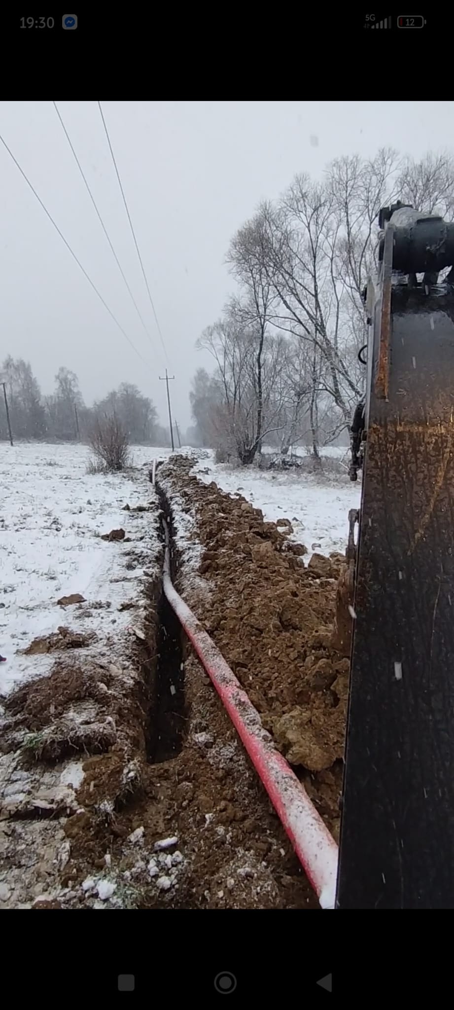 Wykop w zasypanym śniegiem terenie z widoczną czerwoną rurą w ziemi, wykonany prawdopodobnie przy użyciu koparki, której fragment widać po prawej stronie kadru.