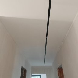 Karol Zep - Wnętrze w trakcie remontu z zamontowaną czarną szyną oświetleniową na suficie, widoczne ślady szpachlowania i niezakończone obramowania drzwi