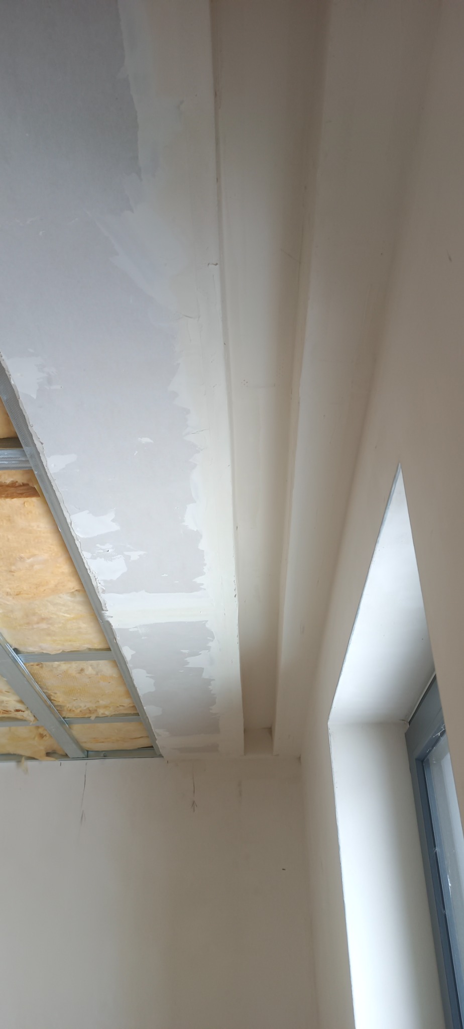 Wnętrze w trakcie prac wykończeniowych: widoczny stelaż sufitu podwieszanego z izolacją, częściowo pokryty płytami gipsowo-kartonowymi, oraz fragment ściany z oknem i nierównościami po nałożeniu...