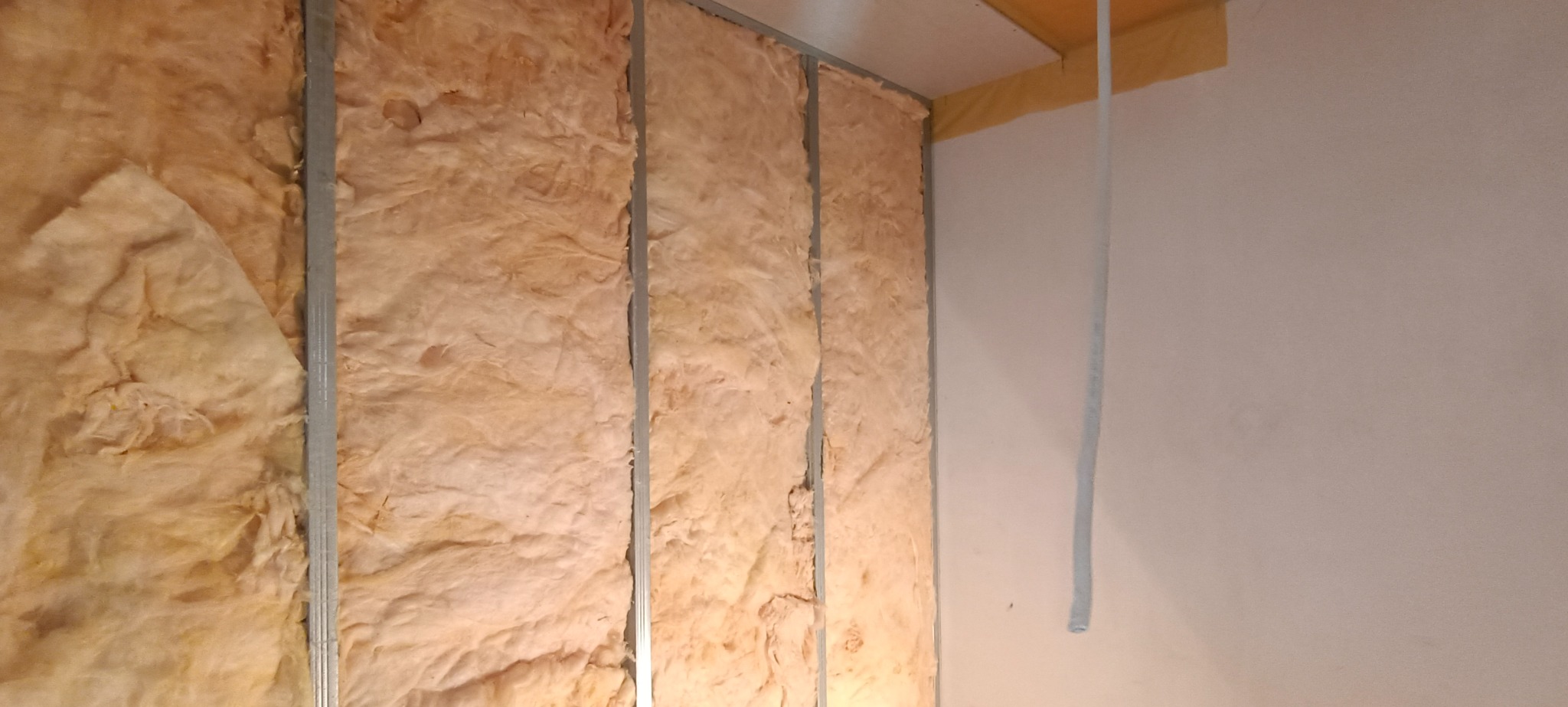 Montaż izolacji z wełny mineralnej w metalowym stelażu na ścianie, widoczny fragment sufitu z taśmą malarską i przewód elektryczny.
