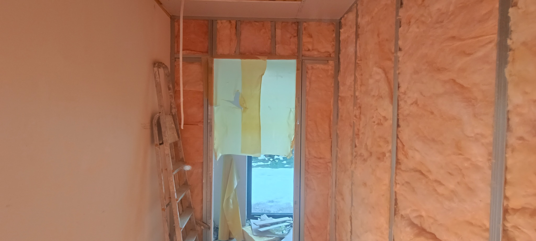 Montaż izolacji z wełny mineralnej w metalowym stelażu ściany działowej, widoczna drabina malarska i zabezpieczone okno folią malarską.