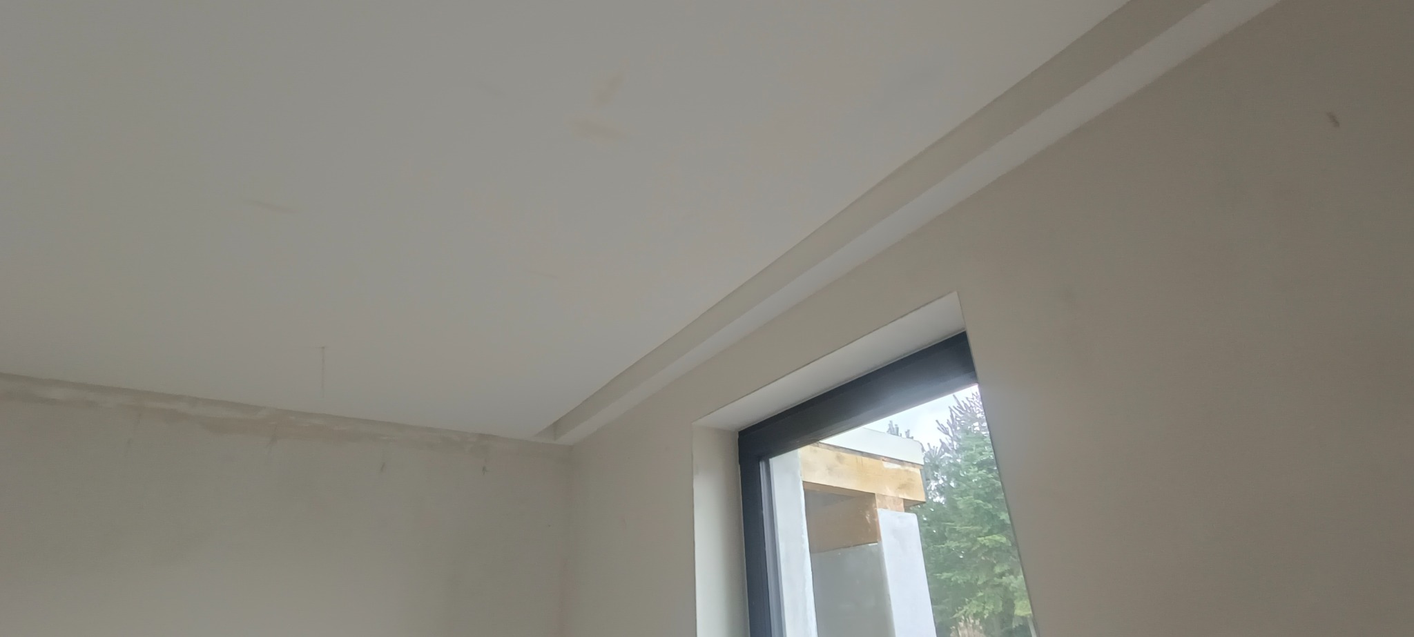 Wykończone wnętrze z pomalowanymi ścianami w jasnym odcieniu i zamontowanym karniszem nad oknem, widoczne niedoskonałości na suficie.