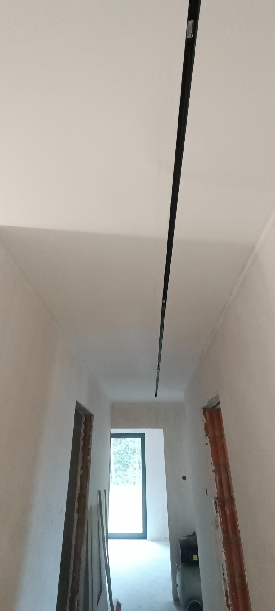 Wnętrze w trakcie remontu z zamontowaną czarną szyną oświetleniową na suficie, widoczne ślady szpachlowania i niezakończone obramowania drzwi
