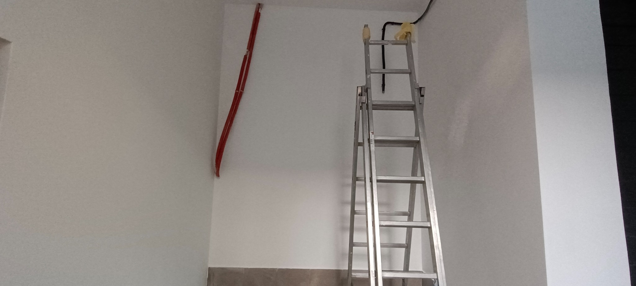 Świeżo pomalowane, białe ściany w rogu pomieszczenia z widocznymi czerwonymi kablami elektrycznymi oraz aluminiowa drabina przystawiona do ściany z czarnym kablem elektrycznym przy suficie.