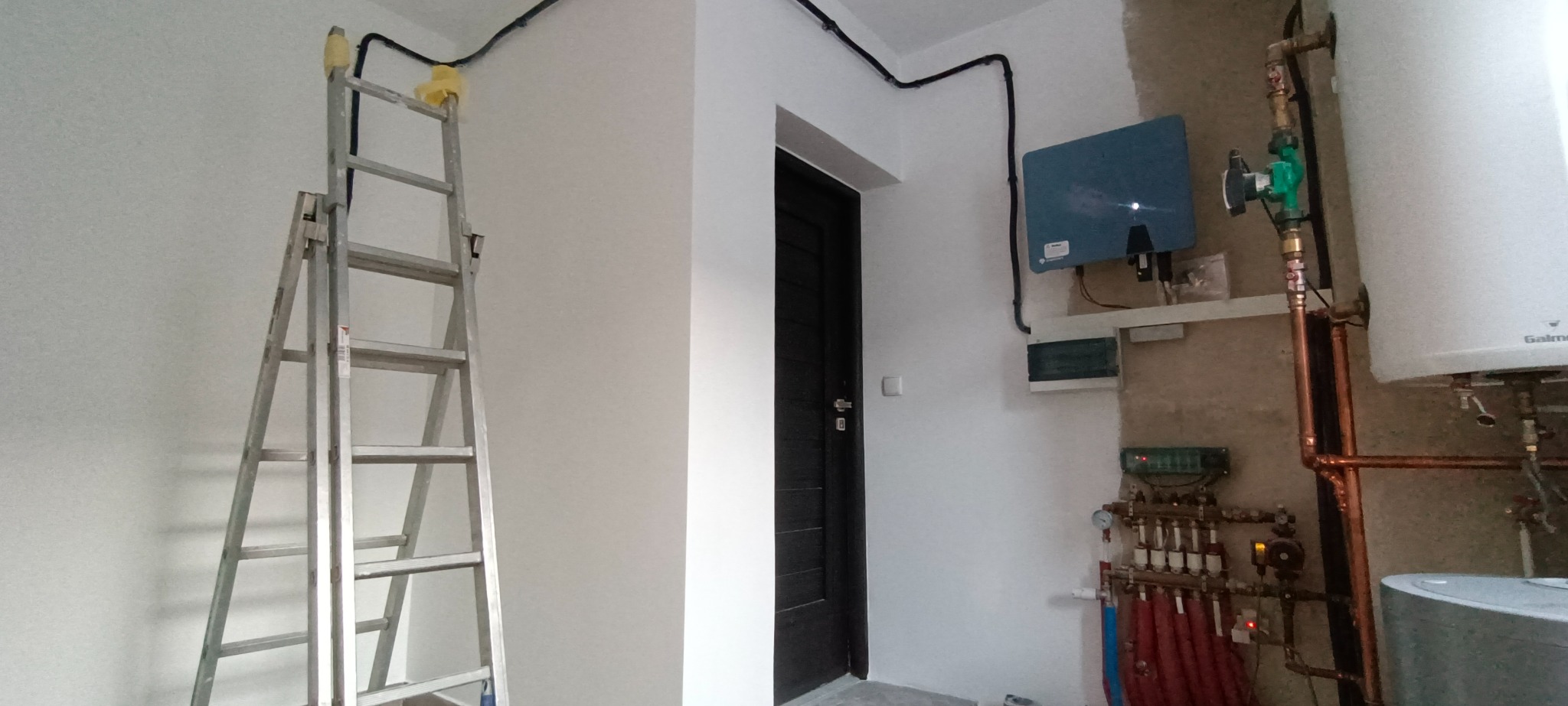 Metalowa drabina oparta o ścianę z poprowadzonymi kablami elektrycznymi, w tle instalacja grzewcza z kotłem i rozdzielaczem, widoczne rury miedziane i podłączenia.