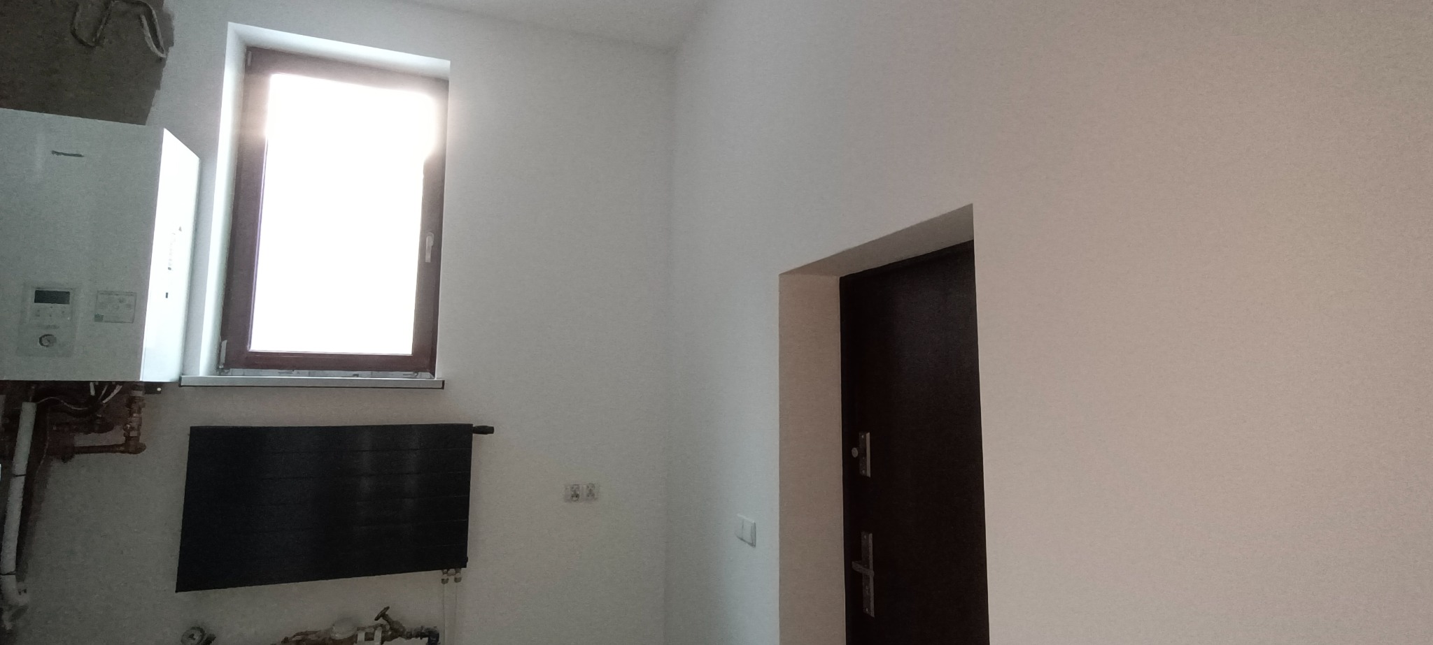 Świeżo pomalowane na biało wnętrze z drzwiami w kolorze ciemnego brązu, grzejnikiem panelowym i kotłem grzewczym, widok na okno z silnym światłem dziennym.