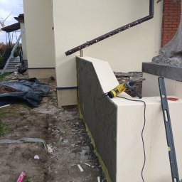 Nazar hulenchin - Ocieplanie elewacji: fragment ściany z nałożoną warstwą kleju na siatce, widoczny styropian, poziomica, wiertarka udarowa oraz fragment rynny i rzeźby w tle.