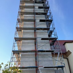 Nazar hulenchin - Wysokie rusztowanie otaczające budynek z elewacją w trakcie prac, widoczne fragmenty cegieł i nałożony szary tynk, fragment dachu z czerwonej dachówki w tle, ujęcie z dołu w słoneczny dzień.