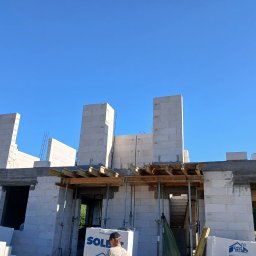 Nazar hulenchin - Budowa domu w trakcie wznoszenia ścian z białych bloczków, widoczne drewniane szalunki i zbrojenia, fragment osoby w dolnej części kadru, błękitne niebo w tle.