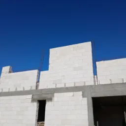 Wznoszenie ścian domu z białych bloczków betonowych, widoczne zbrojenie i otwory okienne na tle błękitnego nieba.