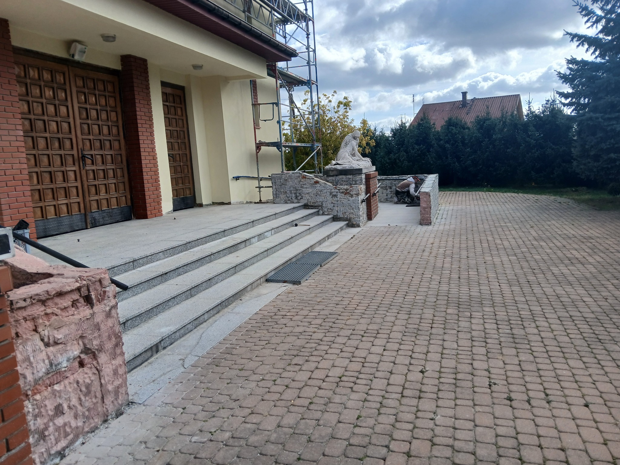 Prace renowacyjne przy wejściu do budynku z kolumnami, widoczne rusztowania, figura dekoracyjna i pracownik wykonujący prace wykończeniowe na zewnątrz.